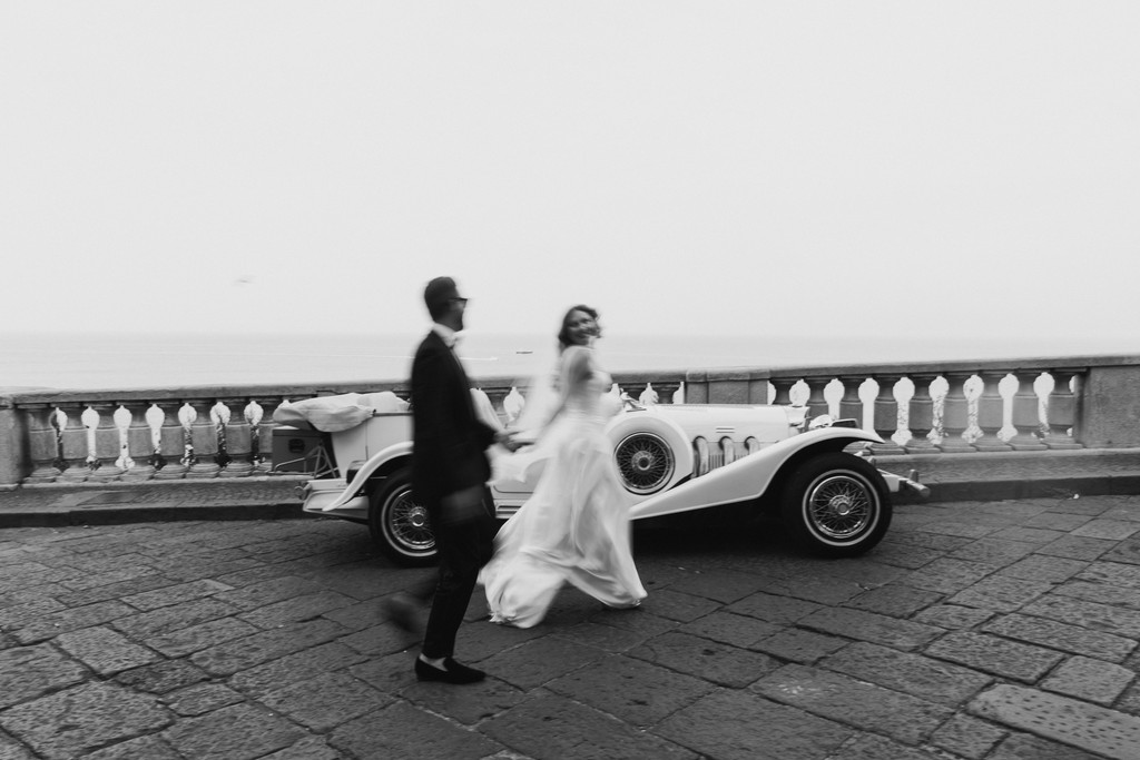     Palm Beach wedding photographer captures a bride and groom walking beside a vintage car in an editorial black and white portrait on the coast of Italy.