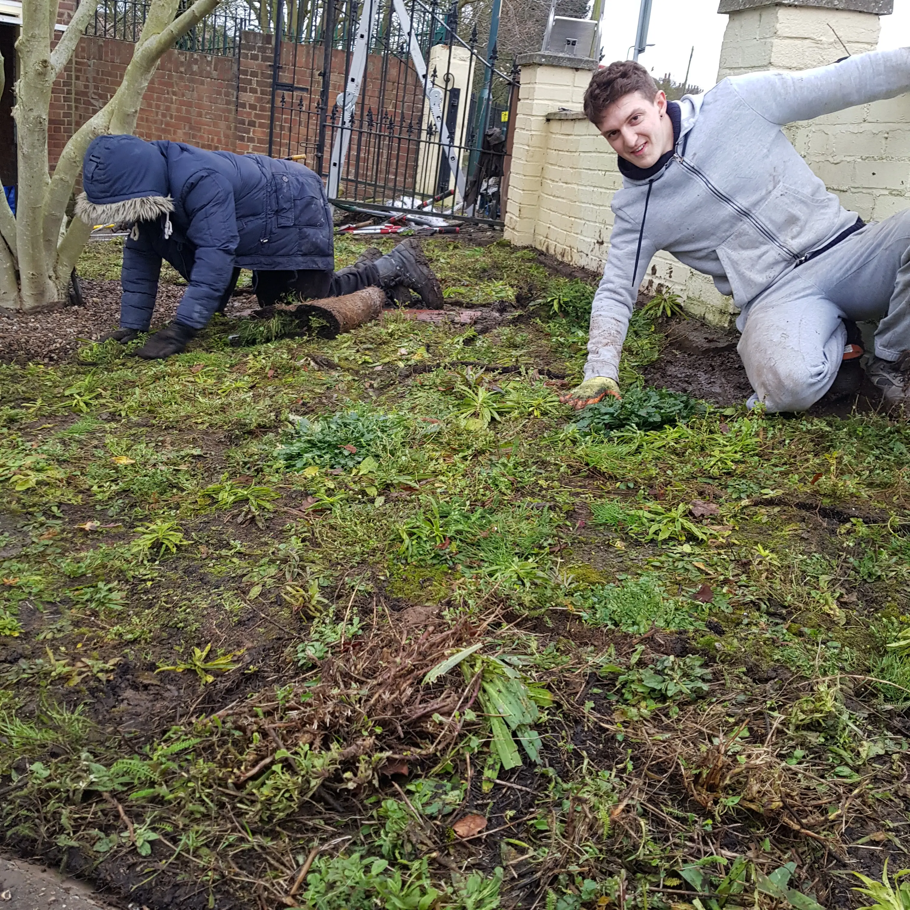 Two people working in a garden, one kneeling and the other posing playfully, surrounded by green grass.
