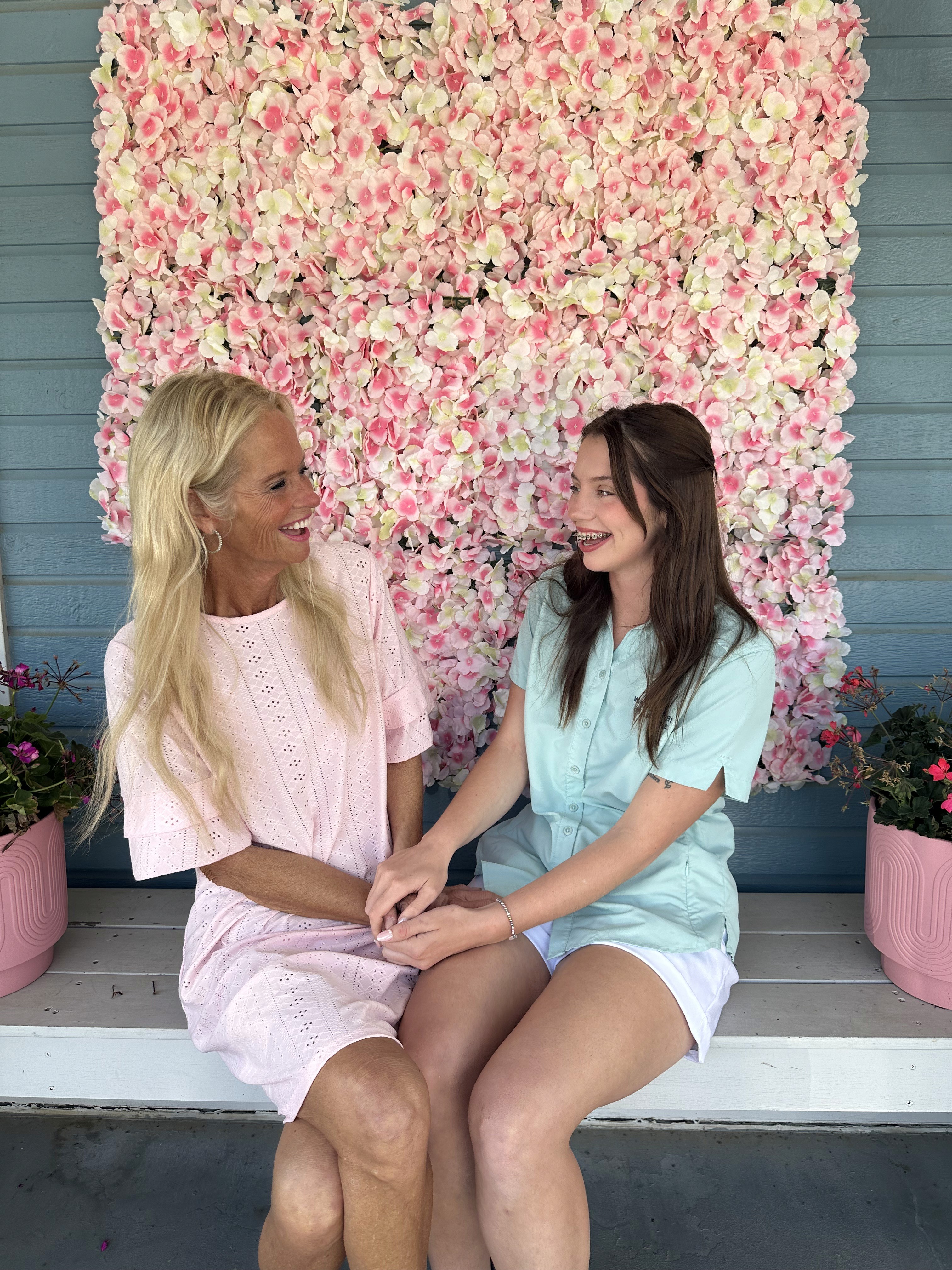 Two women smiling and holding hands, sitting on a porch bench in front of a wall adorned with pink and white flowers, surrounded by potted plants.