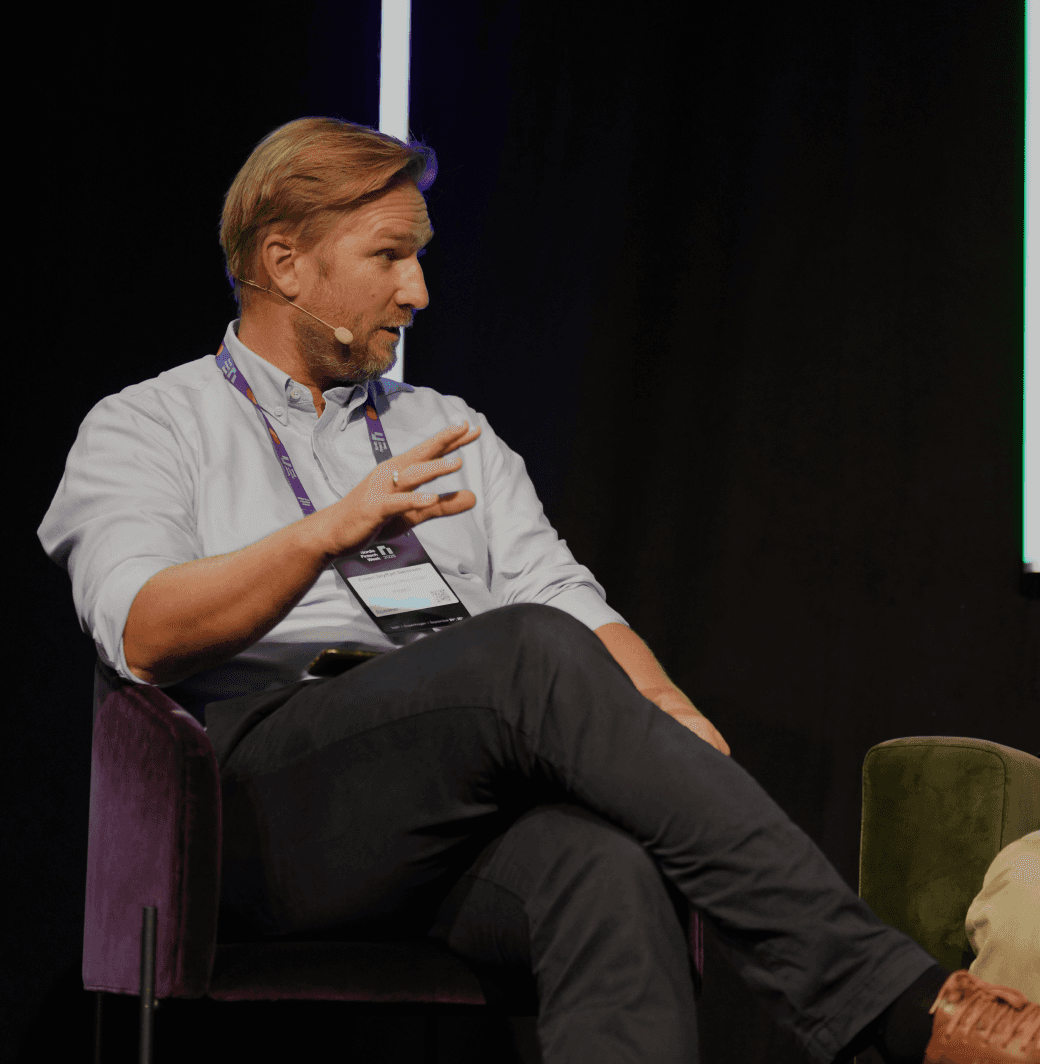 A person sitting on a stage, engaging in discussion, wearing a light shirt and gesturing with one hand.