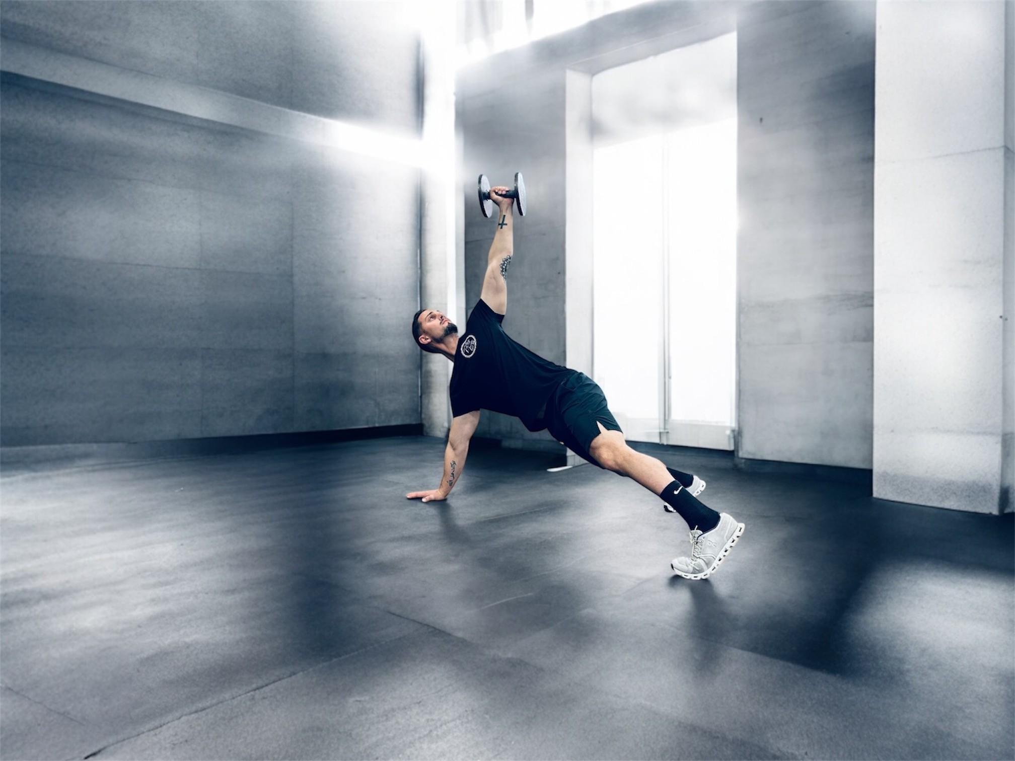 Man performing a dumbbell plank exercise, lifting a weight above his head in gym attire.