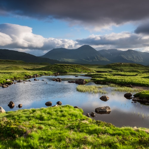 A serene landscape featuring a calm river flowing through lush green fields with rocky outcrops and distant mountains under a cloudy sky.