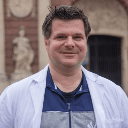 Lächelnder Apotheker mit einem weißen Kittel, der vor einem historischen Gebäude mit verzierten Elementen steht.