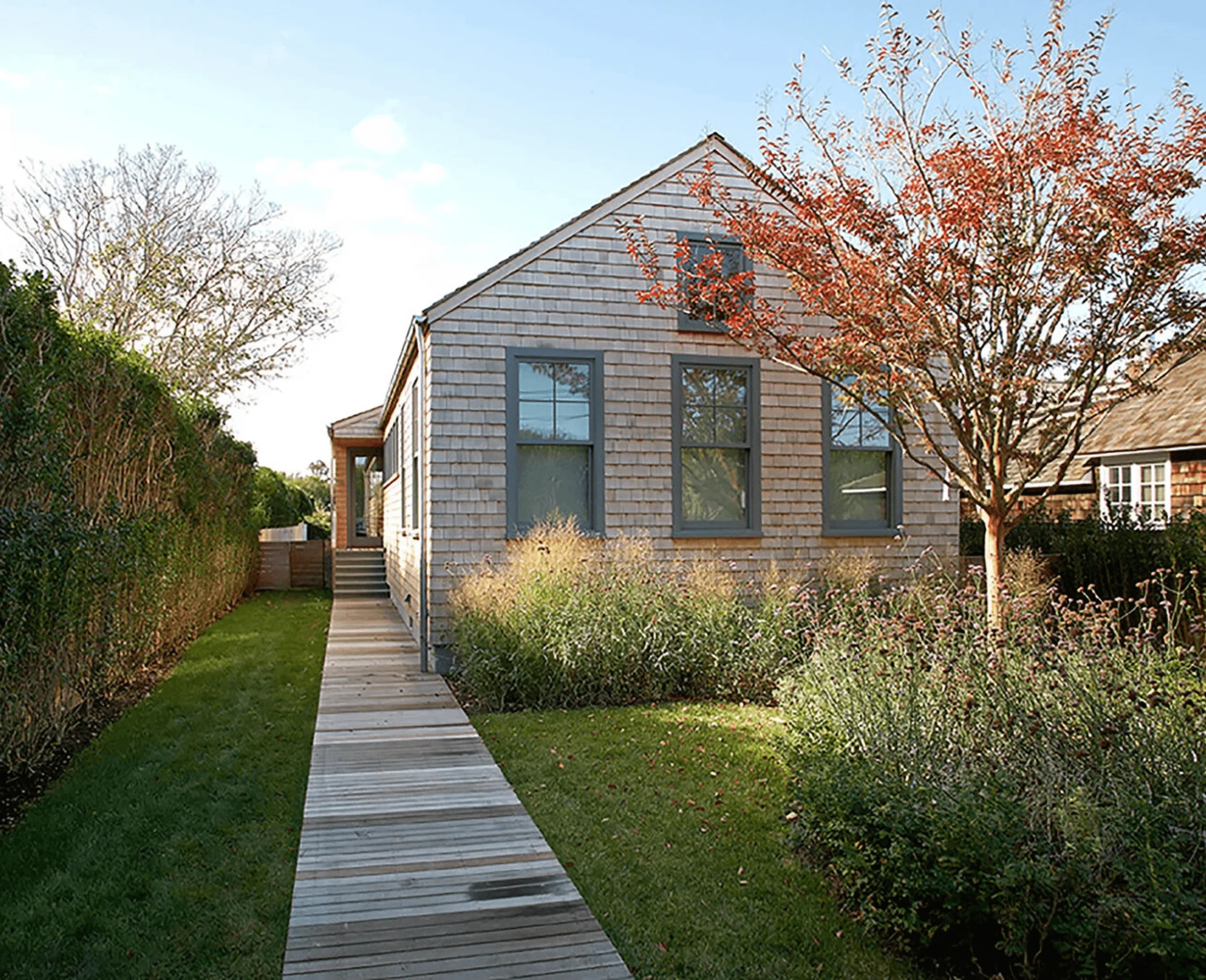 The shingled exterior of the Sagaponack Cottage, with a wood walkway leading through native grasses to the entrance