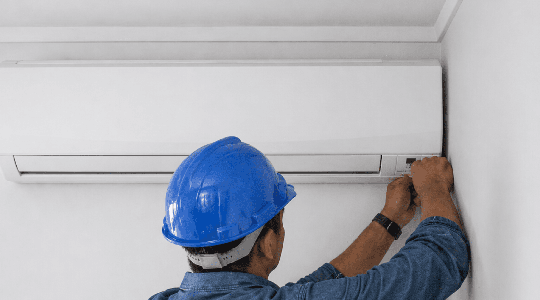 A technician in a blue hard hat adjusting a wall-mounted air conditioner.