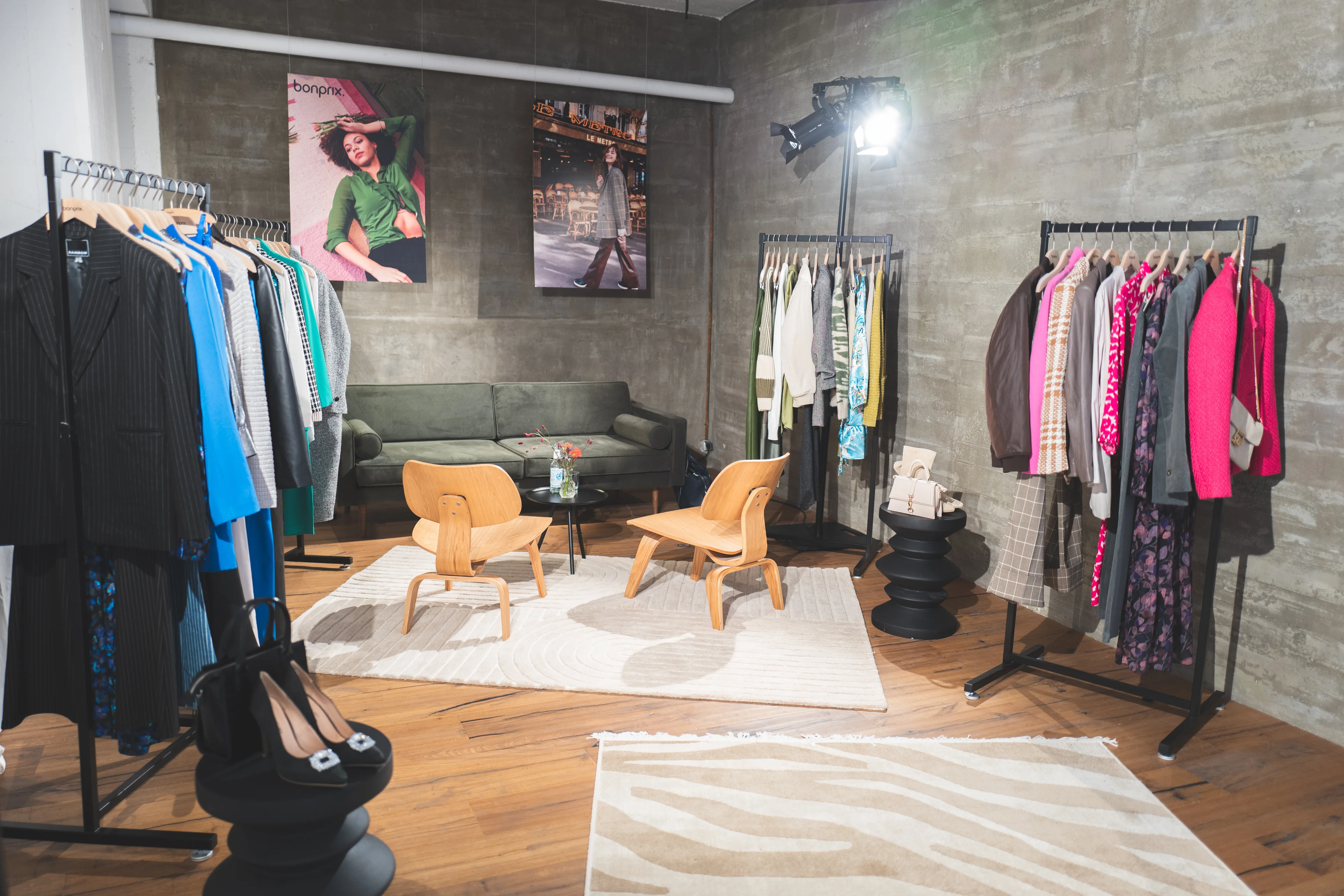 Showroom with clothing racks, green sofa, wooden chairs and Bonprix campaign posters on a concrete wall.
