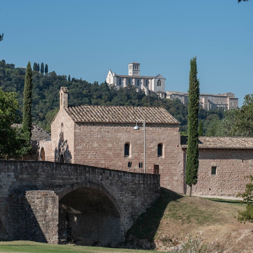 在清澈的蓝天下，有石头建筑、桥梁、高大的柏树，以及背景中的大型山坡修道院。