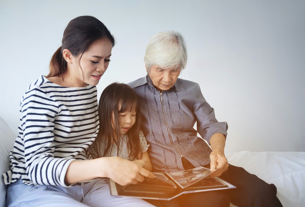 Three generations of an Asian family share a heartfelt moment as they look through a photo album together. The grandmother, mother, and young daughter sit closely, engaged in the memories captured within the album. The scene emphasizes family heritage, storytelling, and the bonding that occurs through sharing family history.