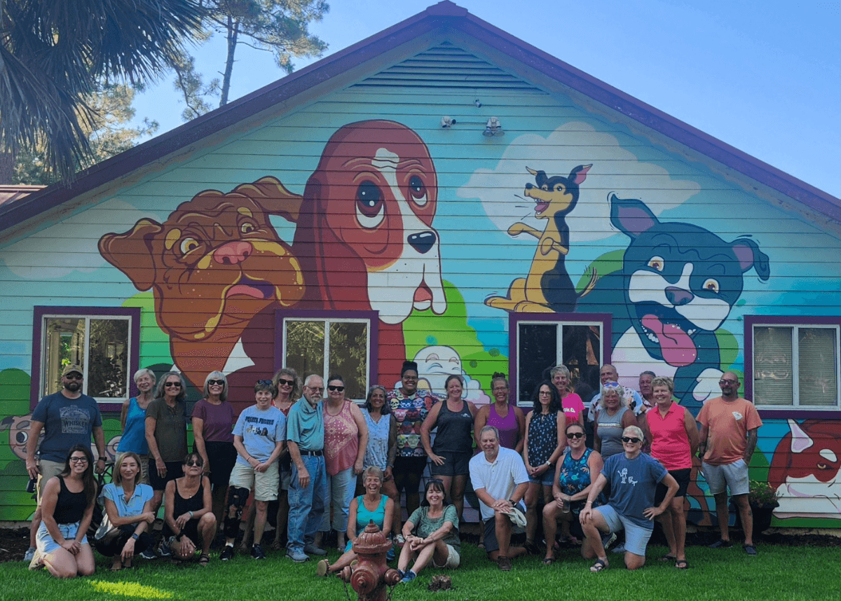 A large group of people stands in front of a colorful mural featuring cartoon dogs and cats.