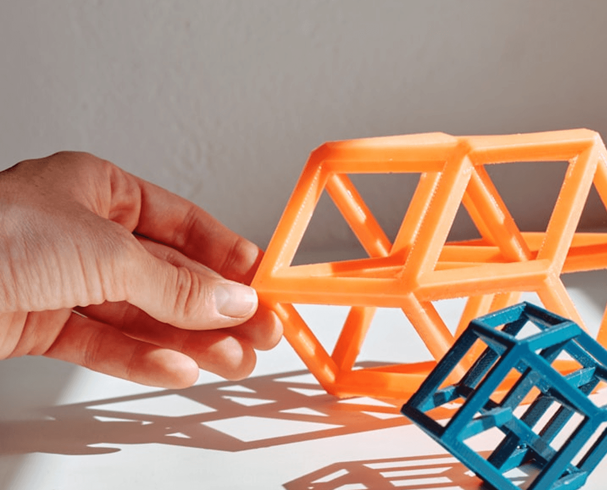 A hand holding a geometric orange 3D-printed structure, resembling interlocking cubes, casting shadows on a white surface beside a similar blue structure; keywords: 3D print, geometric design, modern technology.