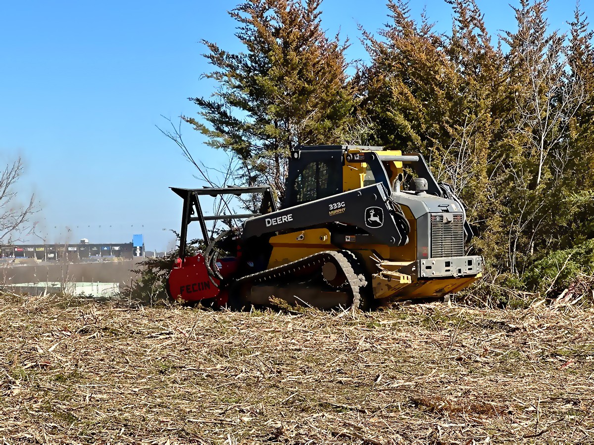 A large yellow excavator is digging a hole in a dirt pile, creating a cloud of dust. The sky is blue with white clouds, and there are trees in the background.