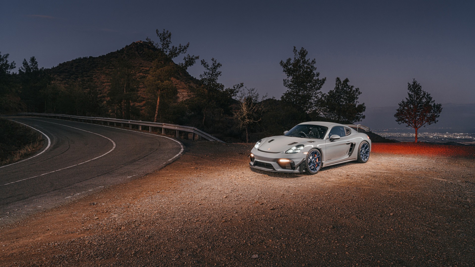 Grey Porsche 718 Cayman GT4 RS with blue wheels parked on a gravel pull-off beside a winding mountain road, headlights on at dusk, with pine-covered hills and a distant city visible below, shot on location in the hills of Cyprus.