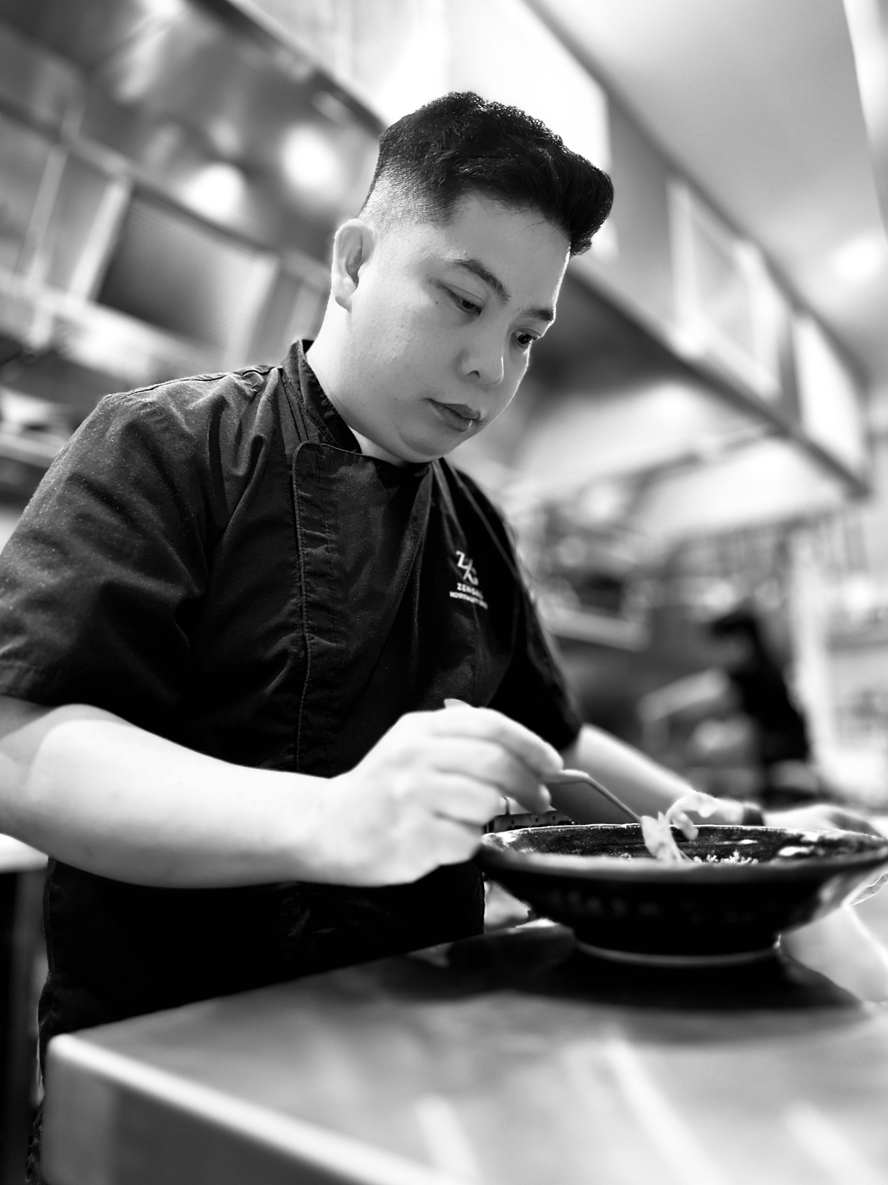 A smiling man with curly hair is sitting at a desk, looking at the camera, with a laptop and a lamp in the background.