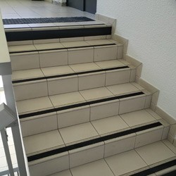 Un couloir carrelé avec un revêtement tactile gris en forme de L près des escaliers et un mur blanc. On aperçoit un escalier avec des rampes noires et une imprimante de bureau.