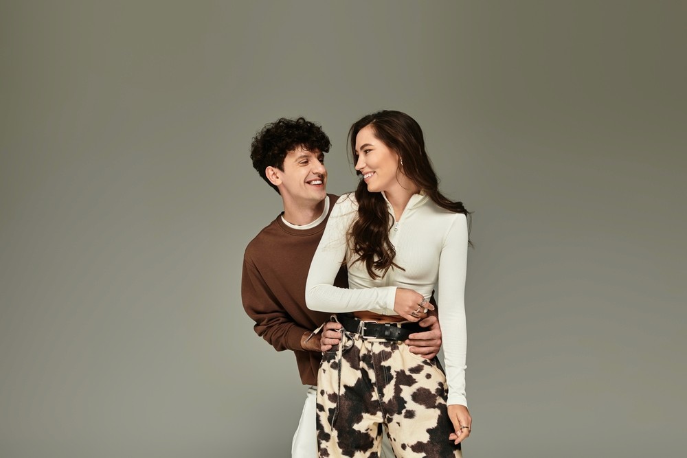 A couple is smiling at each other as the man hugs the woman from behind against a grey background. 