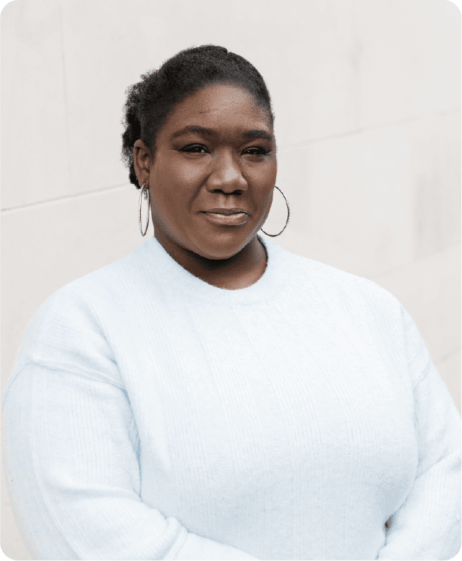 A medium shot captures therapist Sabrina Simpson from the chest up, facing the camera with a slight smile. Sabrina Simpson has dark, curly hair pulled back and wears large, silver hoop earrings. Her light blue, fuzzy sweater has a ribbed texture. The background is a plain, light-colored wall with subtle horizontal lines.