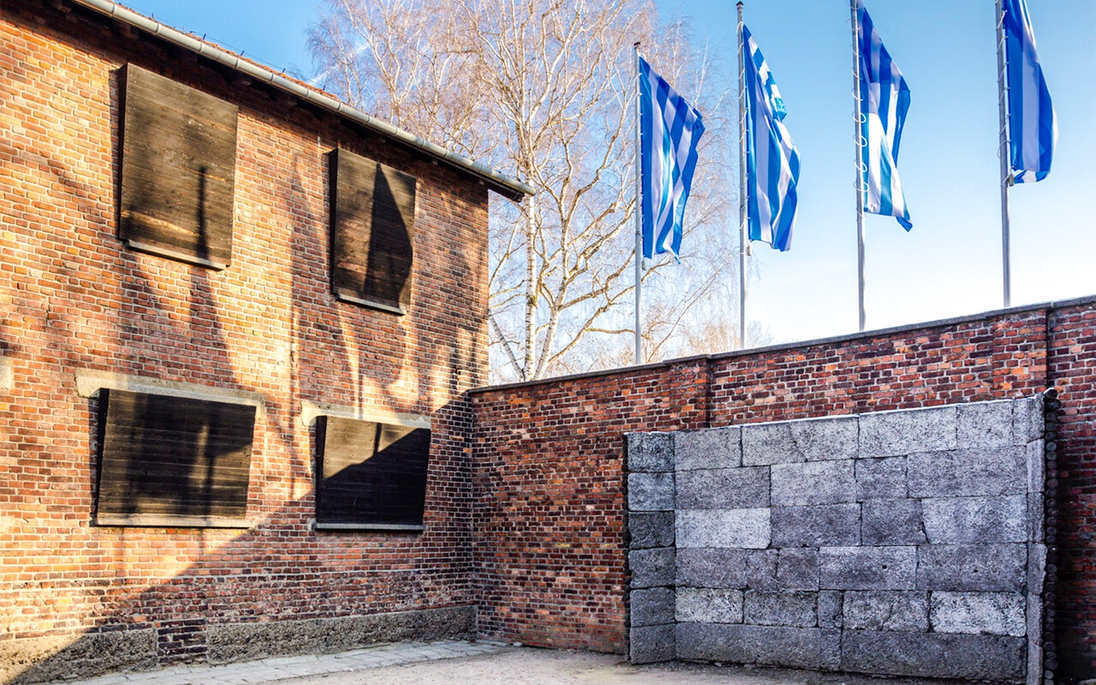 Minnisveggur við Auschwitz-Birkenau búðirnar með múrsteinabyggingu og fánum í bakgrunni.
