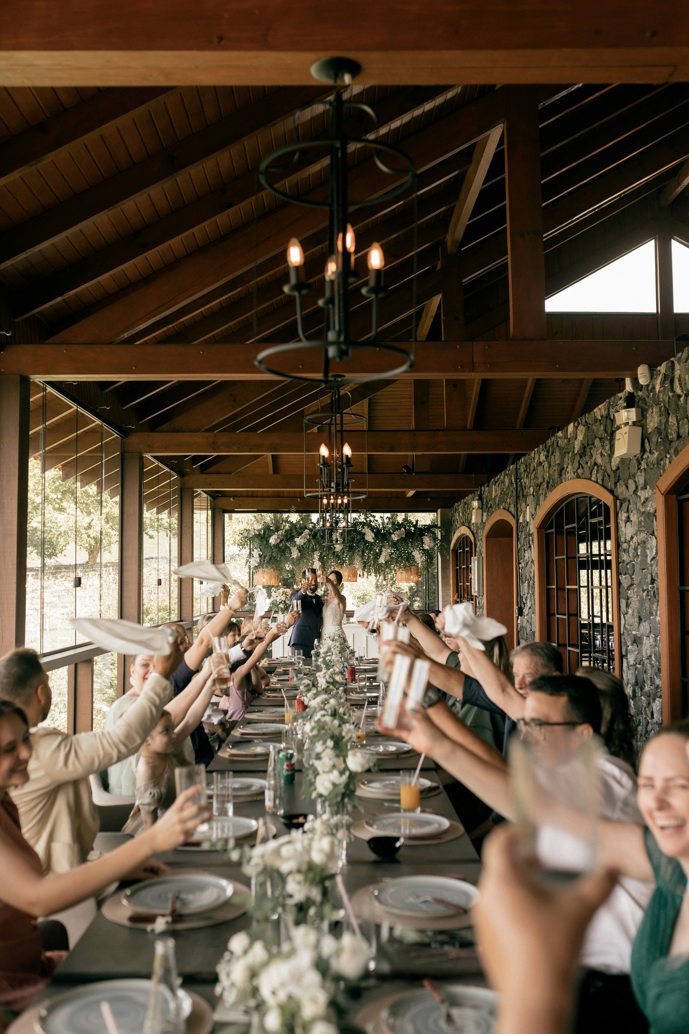 Wedding guests raising glasses and napkins in a toast at a long reception table under a rustic wood-beam pavilion
