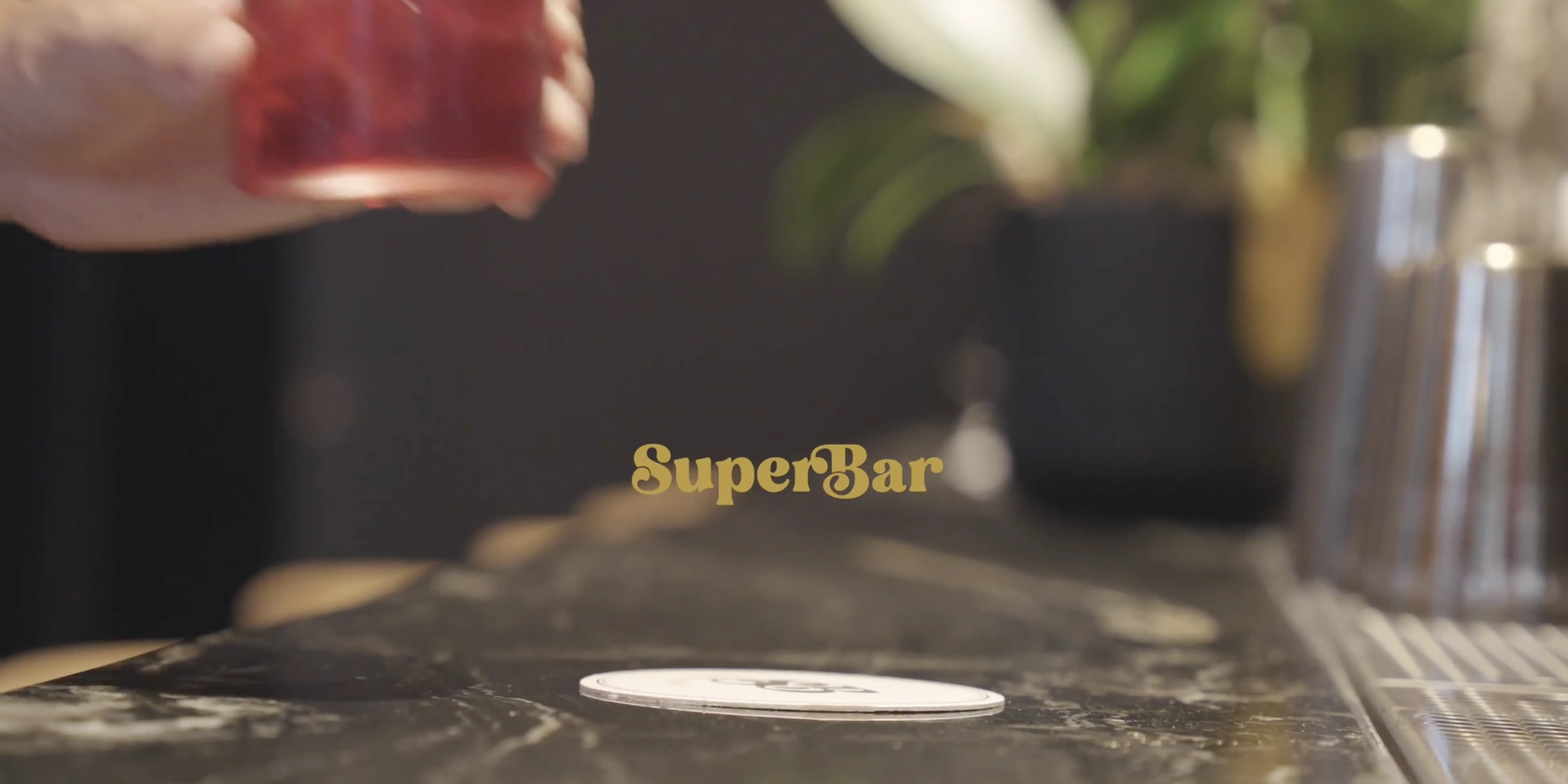 A hand holds a glass filled with a red beverage over a marble counter, with the blurred interior of a modern bar and a coaster with the logo "SuperBar" in focus.