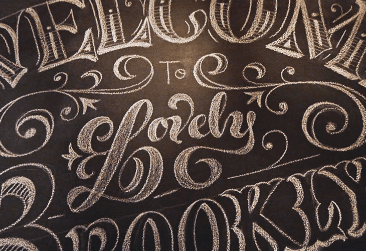 The image shows a close-up of a dark surface, possibly a chalkboard or dark-colored paper, with white cursive text written on it. The text reads "Welcome to Emily," with decorative flourishes and swirls surrounding the words. The style of the lettering is elegant and flowing. The surface appears slightly textured, and the lighting creates subtle highlights and shadows, giving it a three-dimensional appearance.