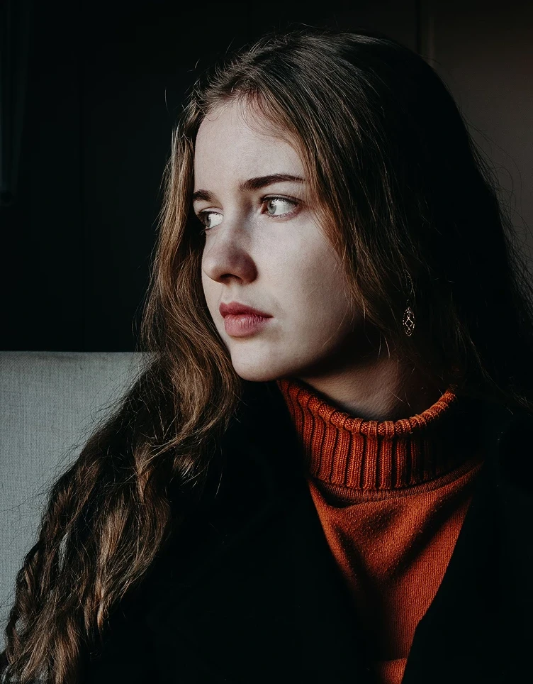 Young woman with long brown hair gazes pensively to the side. She wears an orange turtleneck under a dark coat, contrasting against a dark background.