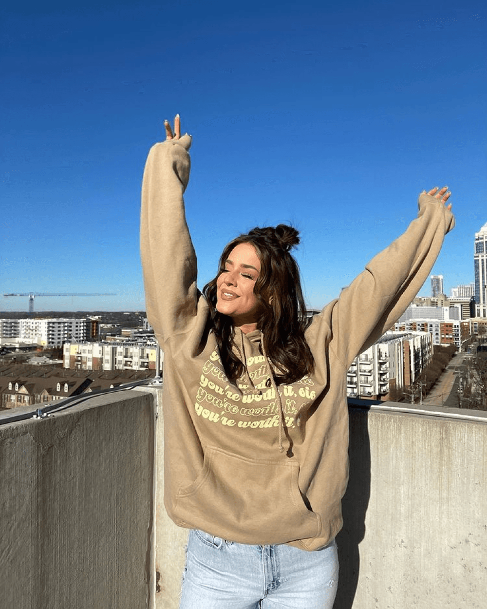 A young woman with brown hair in a tan hoodie stands on a rooftop with her arms raised. A city skyline is in the background.