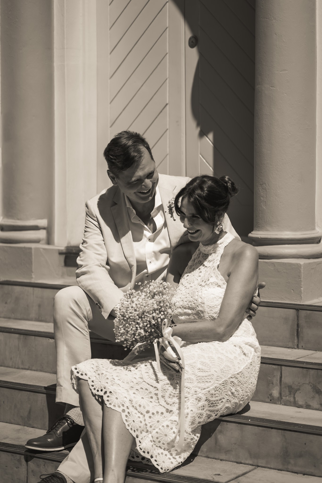 Black and white portrait of a bride and groom seated together on steps beside tall columns.