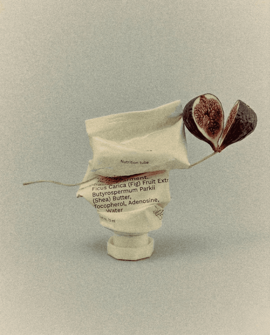 A crumpled handcream in a tube labeled "Nutrition tube" with ingredients text sits upright. A delicate fig branch extends, showcasing a halved fig, adding a surreal, organic touch.
