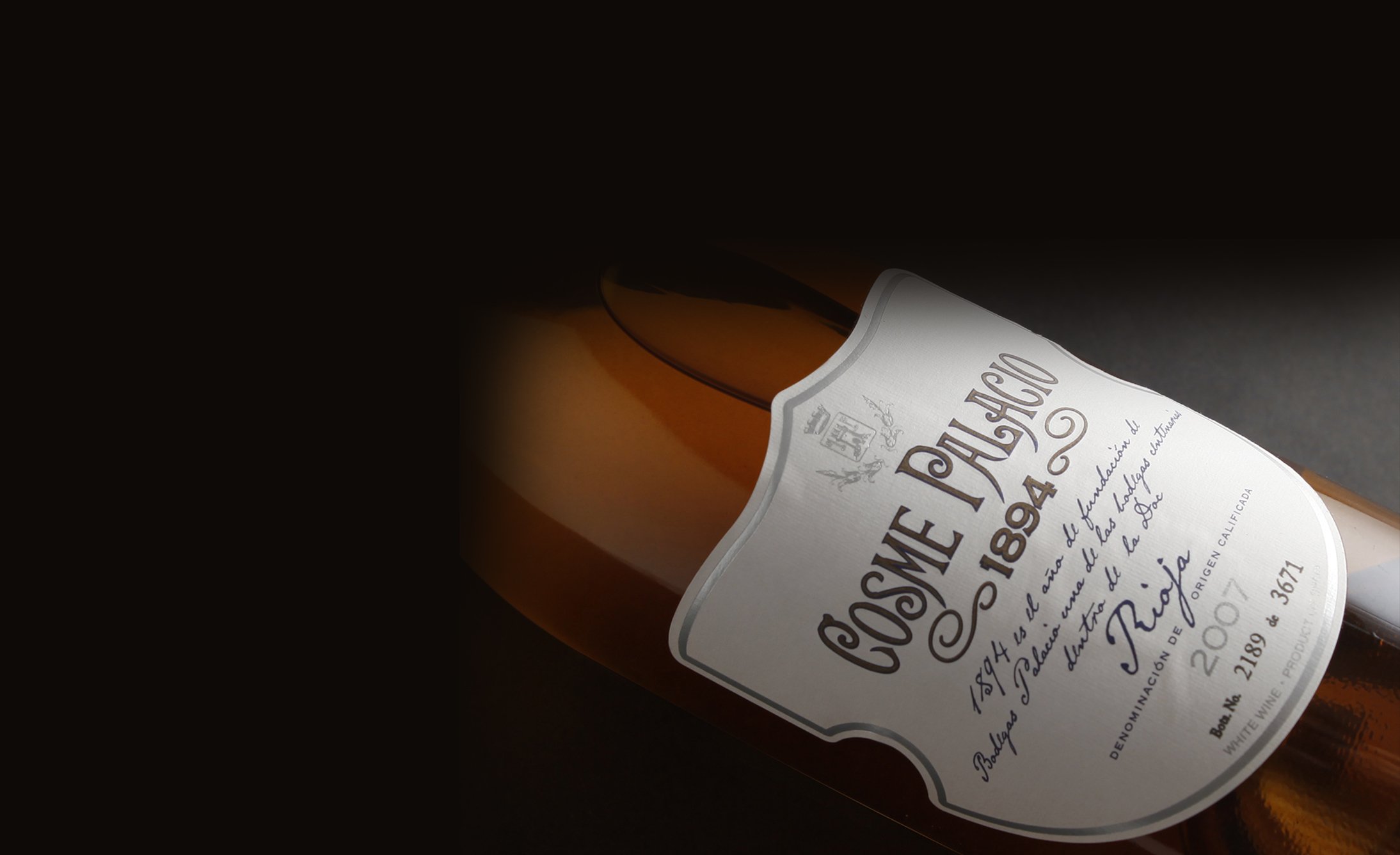 Una botella de vino con una etiqueta blanca que dice "Cosme Palacio 1894" yace de lado. El enfoque está en la elegante escritura y los detalles, establecidos contra un fondo oscuro.