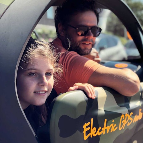 Seorang pria yang memakai kacamata hitam dan seorang anak melihat keluar dari jendela kendaraan bermotif kamuflase dengan tulisan "Electric GPS" di atasnya.