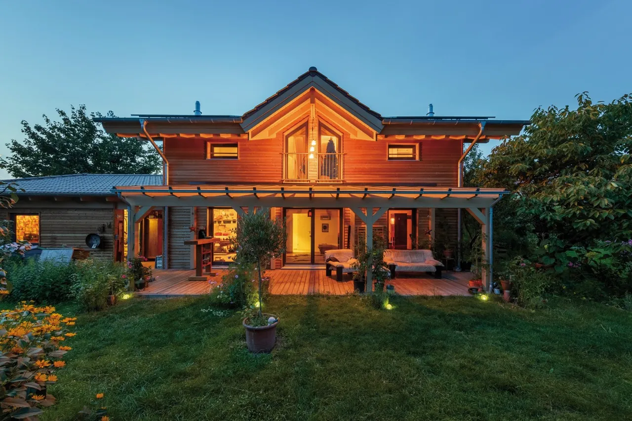 Holzhaus mit Veranda und Garten bei Dämmerung, von außen beleuchtet.