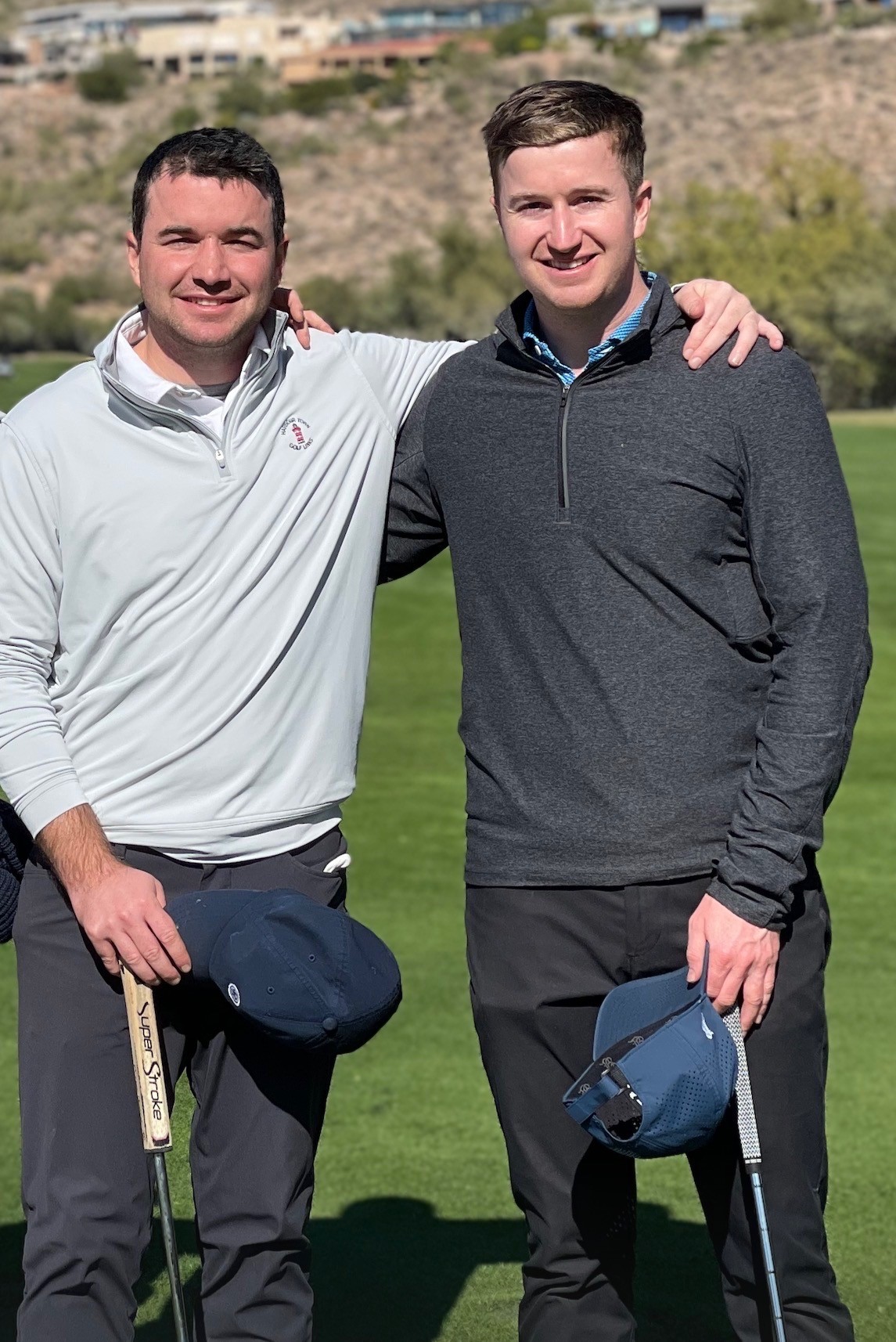 A smiling man with curly hair is sitting at a desk, looking at the camera, with a laptop and a lamp in the background.