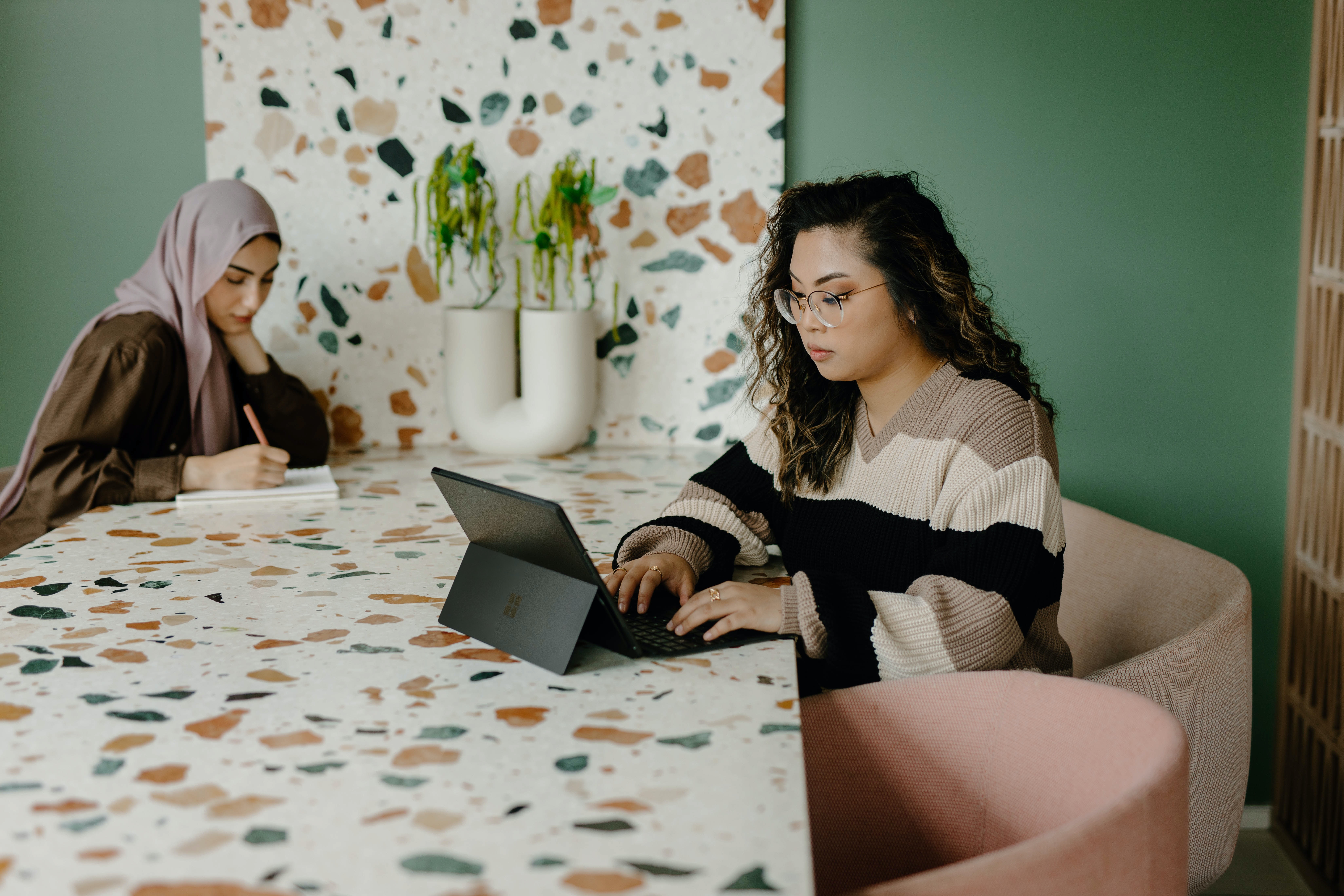 An individual is shown working on a laptop at a table with a patterned surface. The setting might suggest a casual or informal work environment.