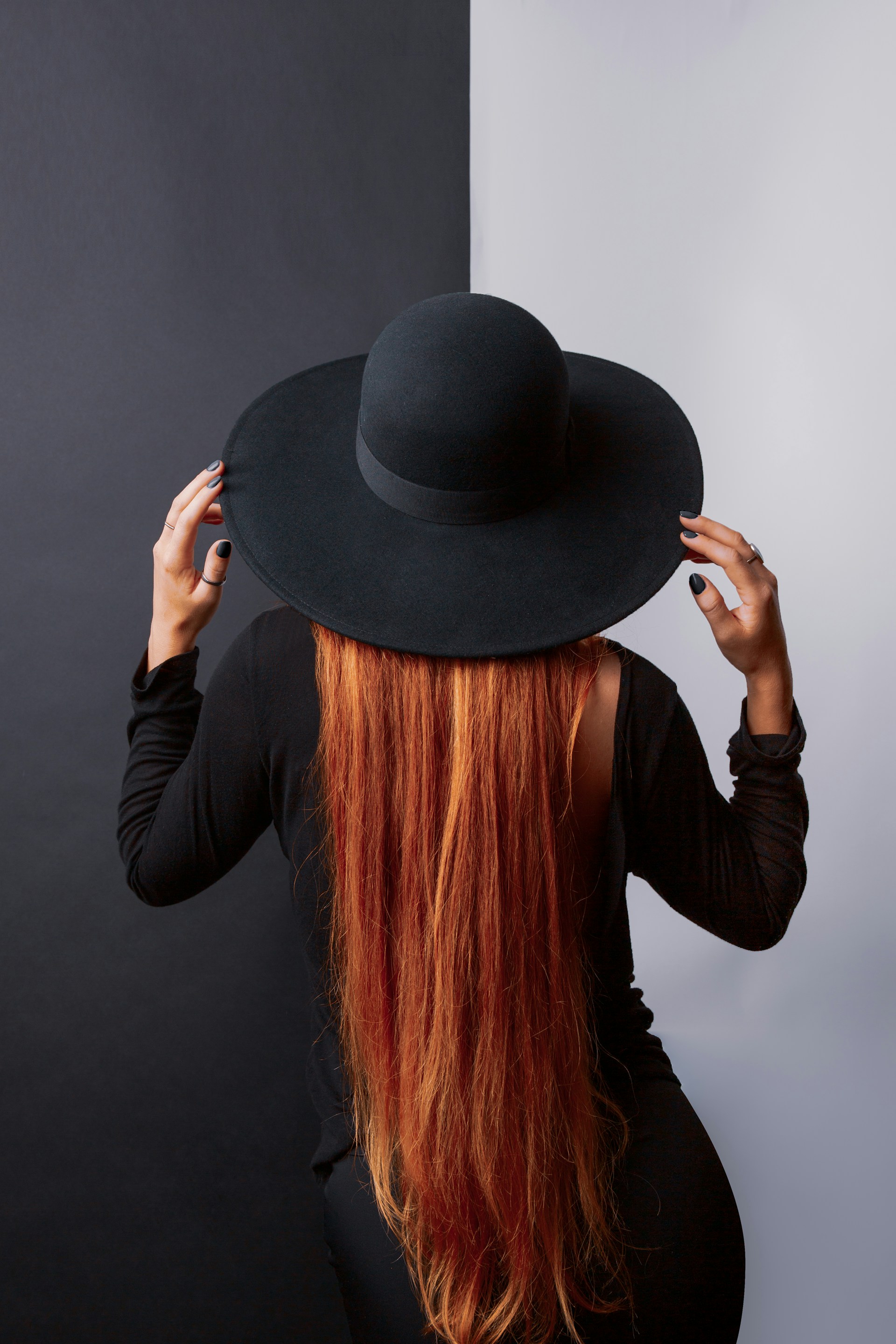 A back view of a woman with long ginger hair and a black wide-brimmed hat. She is wearing a backless black dress and holding the brim of her hat with both hands, featuring black nail polish.