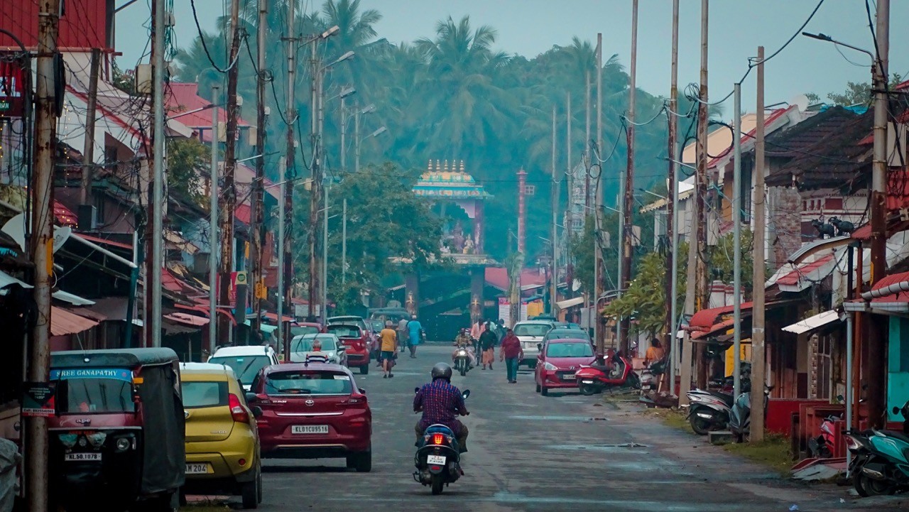 A bustling street in Kalpathi Heritage Village, lined with traditional houses, parked cars, and electric poles. A motorcyclist rides down the road toward a temple with a beautifully decorated entrance, surrounded by coconut trees and misty air. Located 13 km away from Au Revoir Resort.