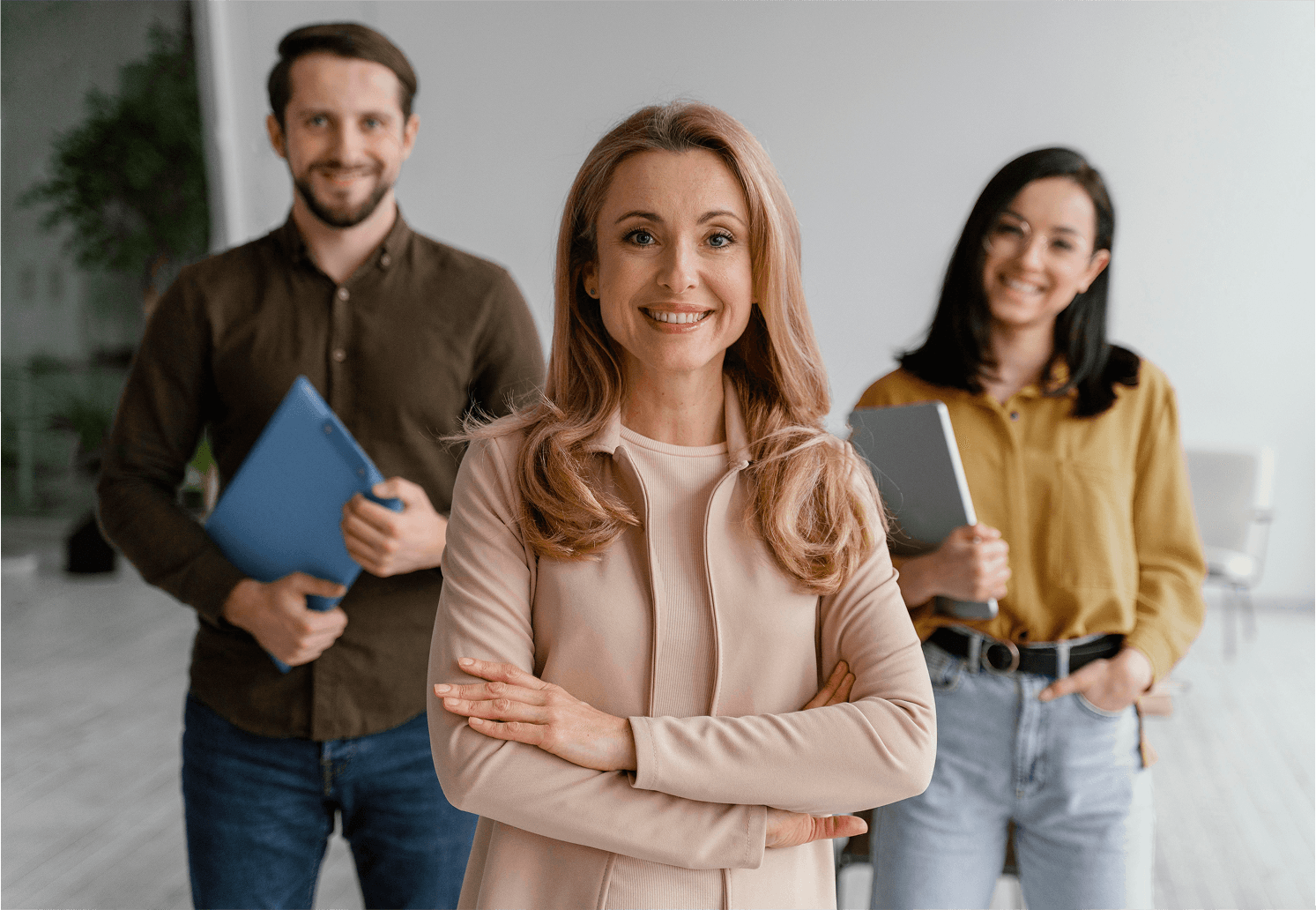 In a modern office setting, three professionals stand confidently; a woman in front wearing a light blazer with her arms crossed, flanked by a man in a brown shirt holding a blue folder and a woman in a mustard blouse holding a gray tablet.