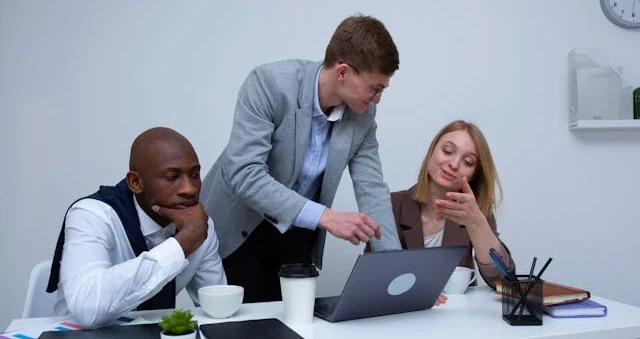 Três colegas em volta de um laptop numa sala clara, um deles de pé