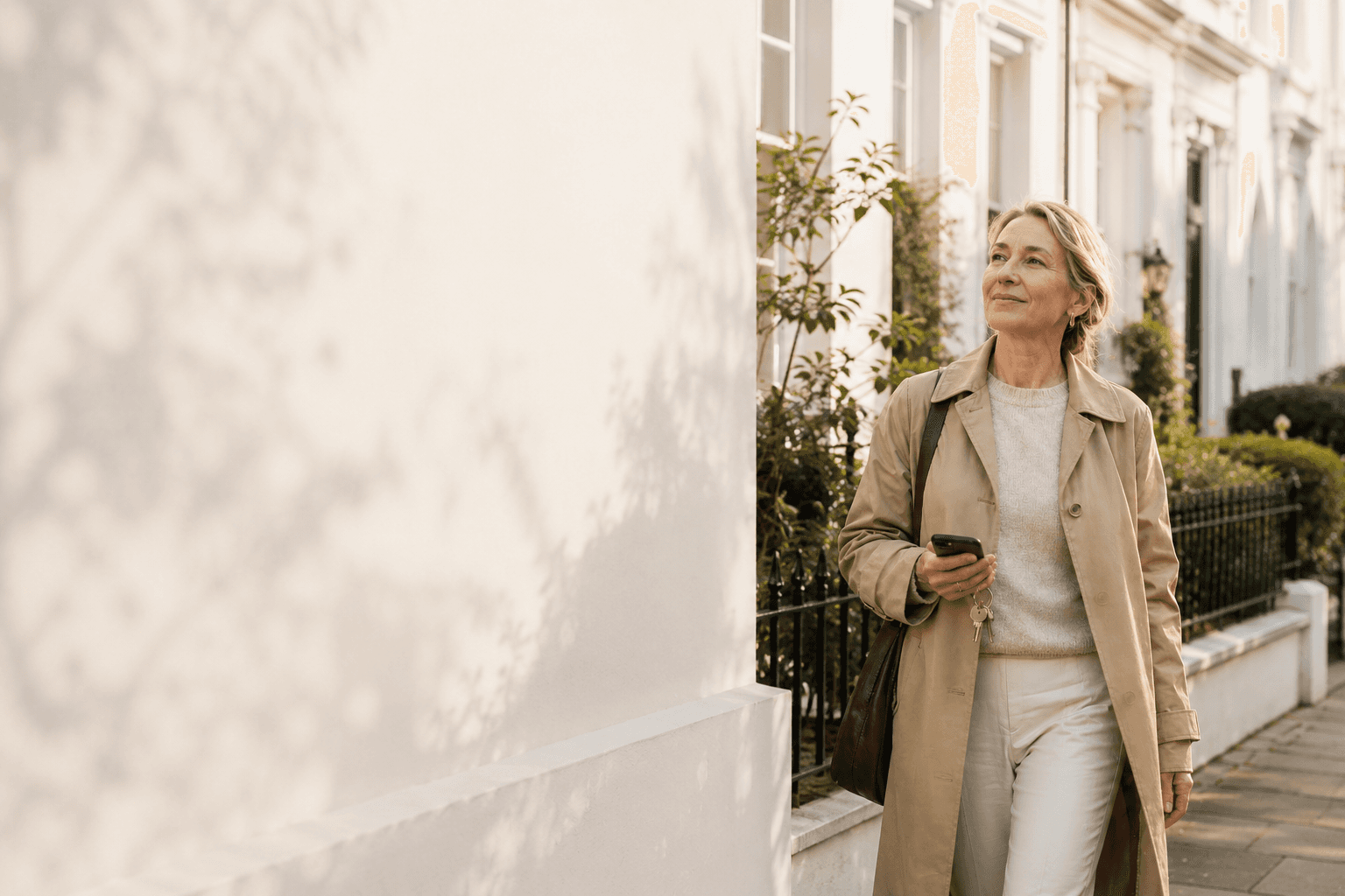 A self-managing accidental landlord checks her phone outside her UK terraced property, holding the front-door keys, in warm afternoon spring light.