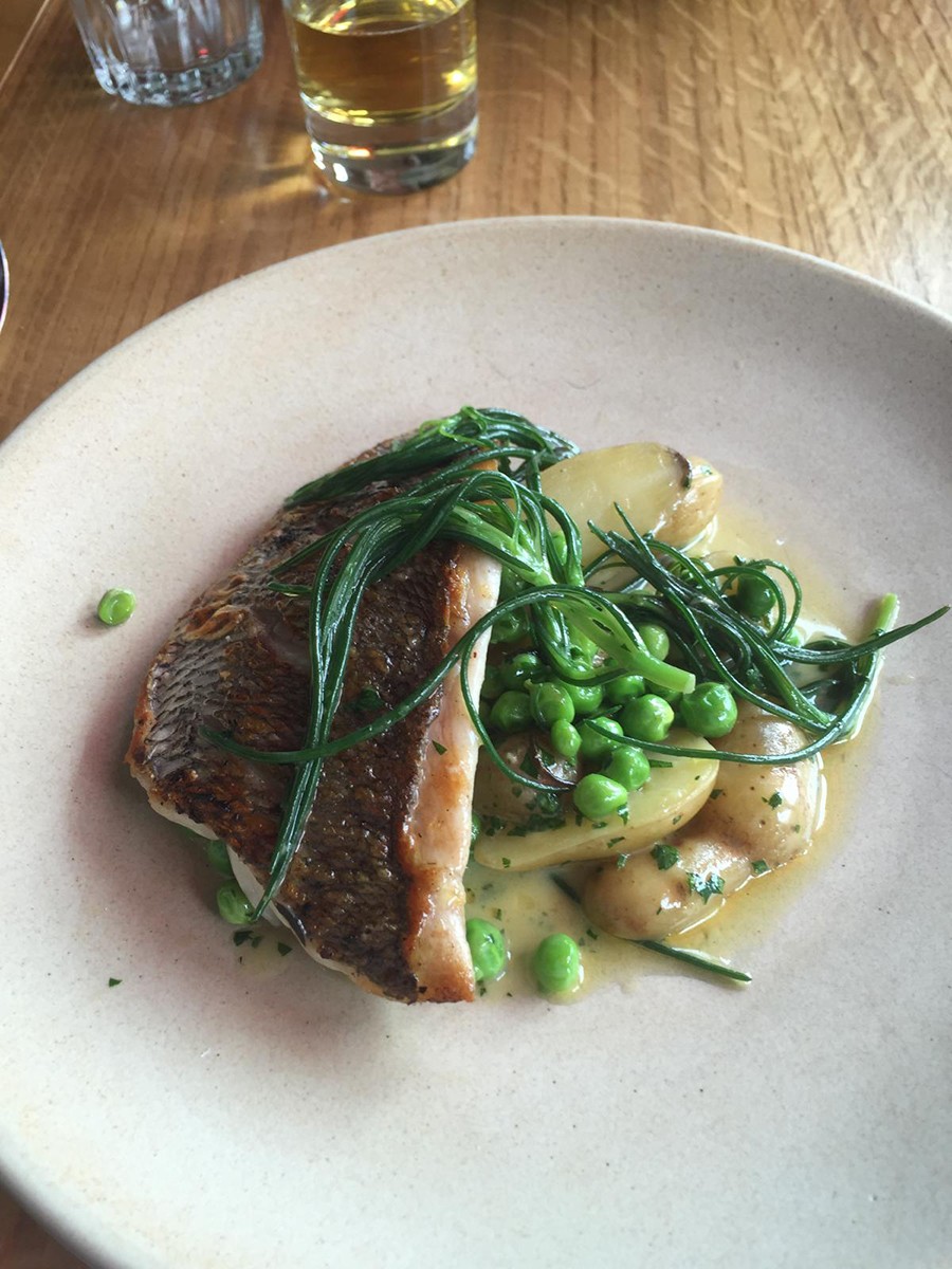 A pan-fried fish fillet served with fresh peas, new potatoes, and fine green herb garnish in a butter sauce, on a ceramic plate.