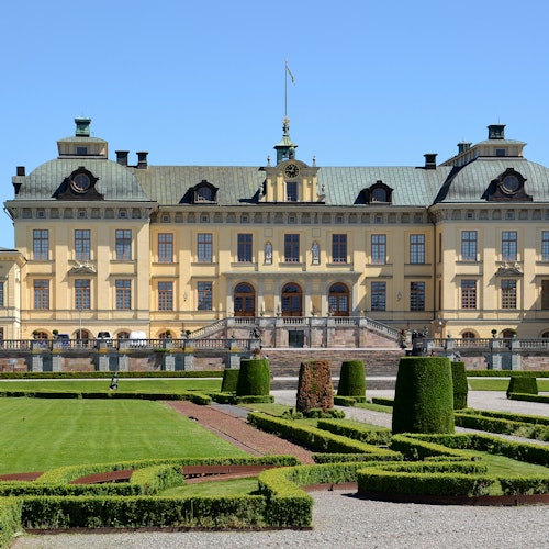 Nagyszerű, sárga barokk palota szimmetrikus ablakokkal, díszes tetőkkel, központi lépcsőházzal és gondozott kertekkel az előtérben.