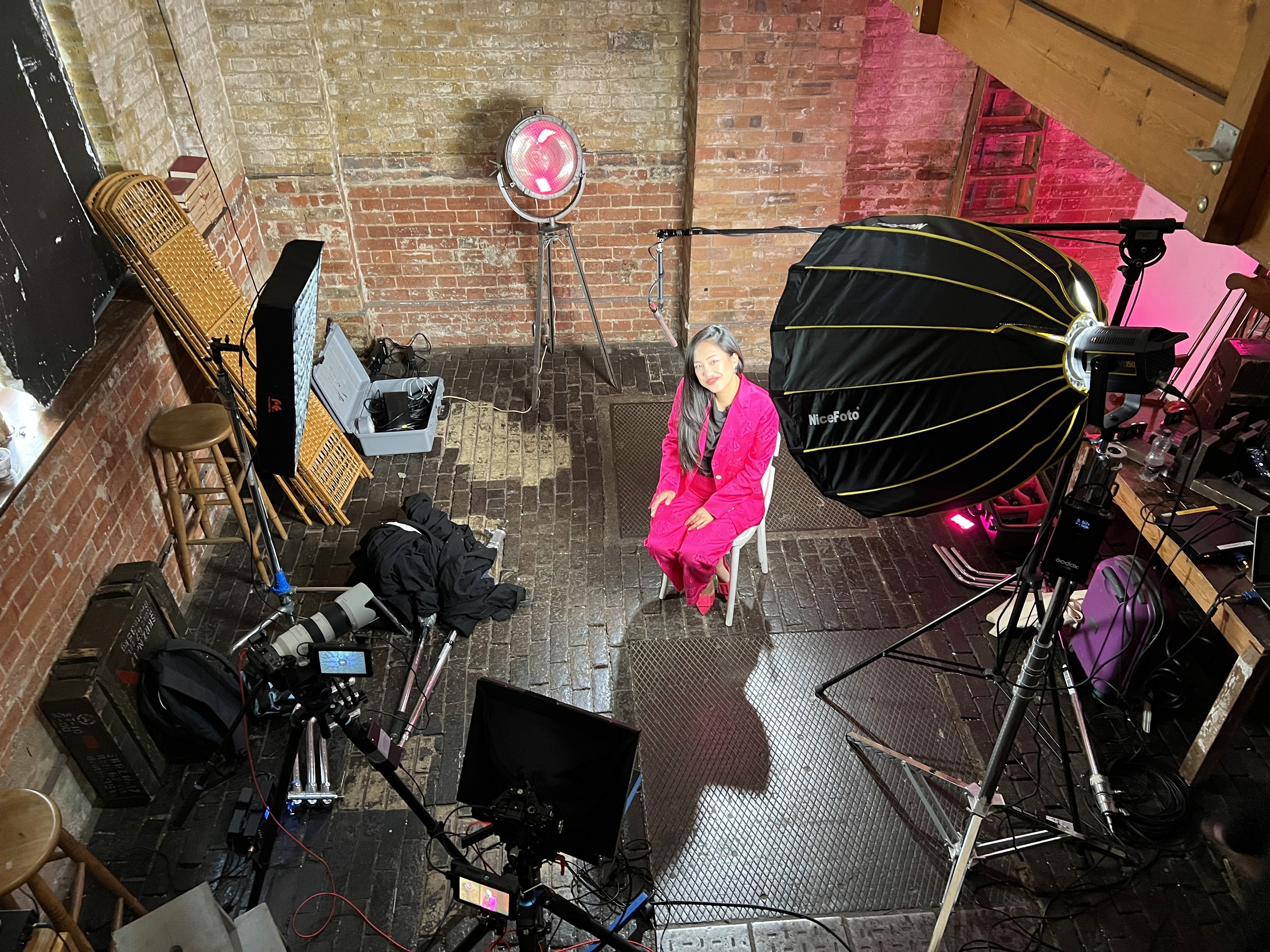 A woman with short brown hair, wearing a floral orange dress, sits at a news desk. She looks serious. Blurred cityscape and cameras surround her.