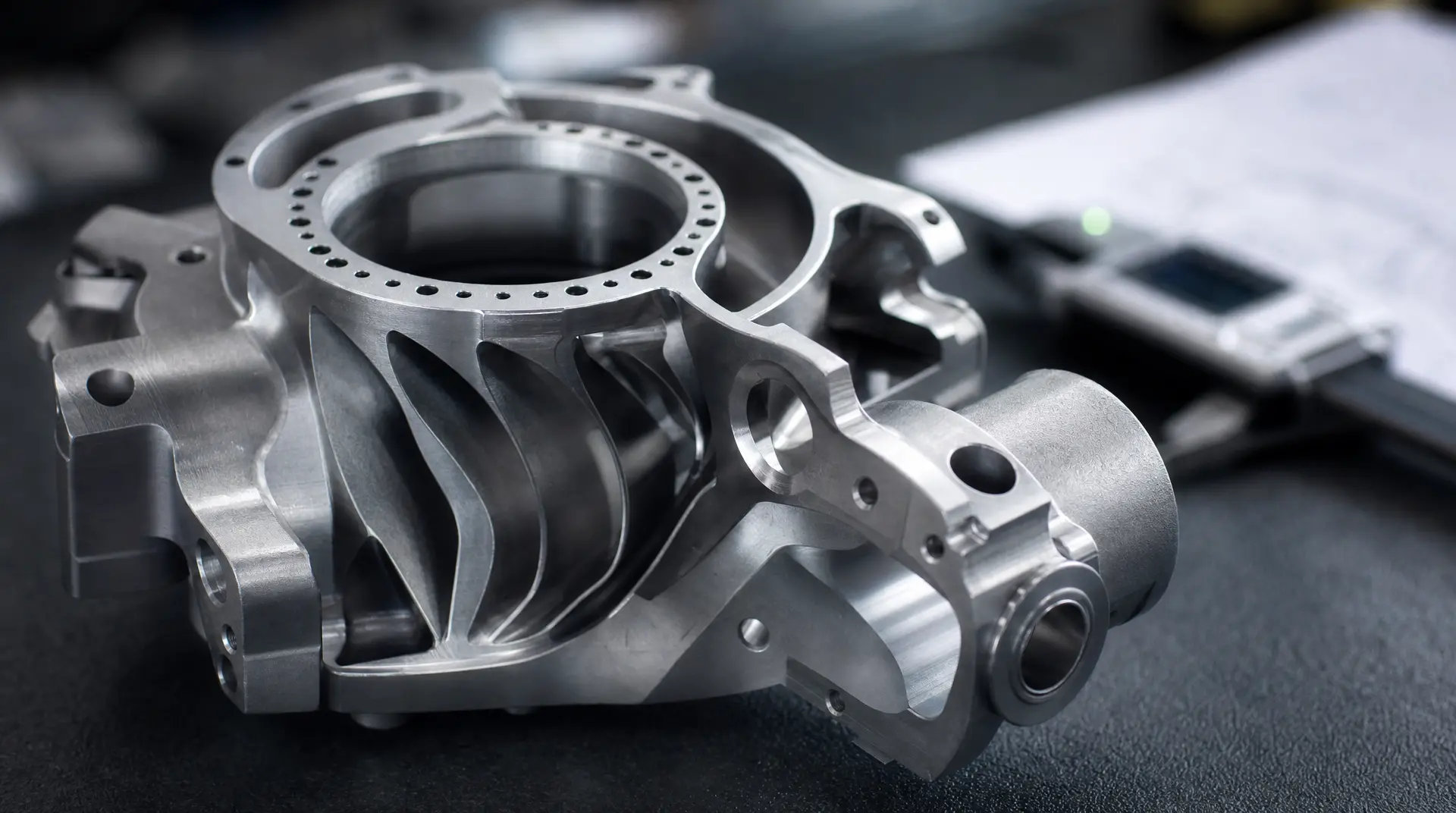 Close-up of a brushed-metal turbine housing with circular openings and machined fins, shot on a dark studio surface with shallow depth of field, aligned to manufacturing video marketing messaging.