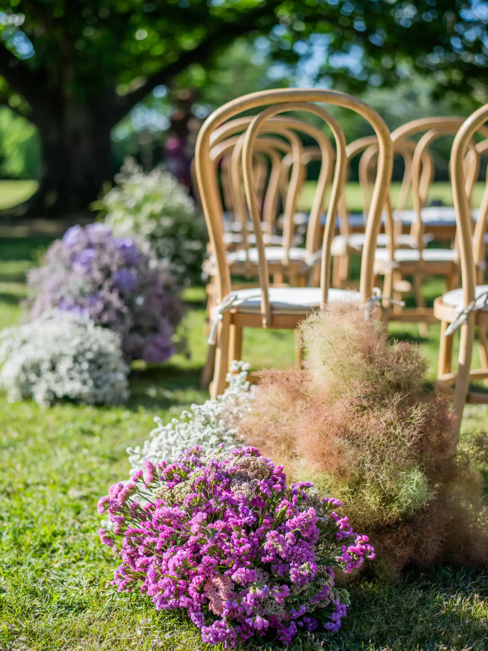 Pasillo de ceremonia al aire libre con filas de sillas de madera y arreglos florales en tonos lilas, blancos y verdes.