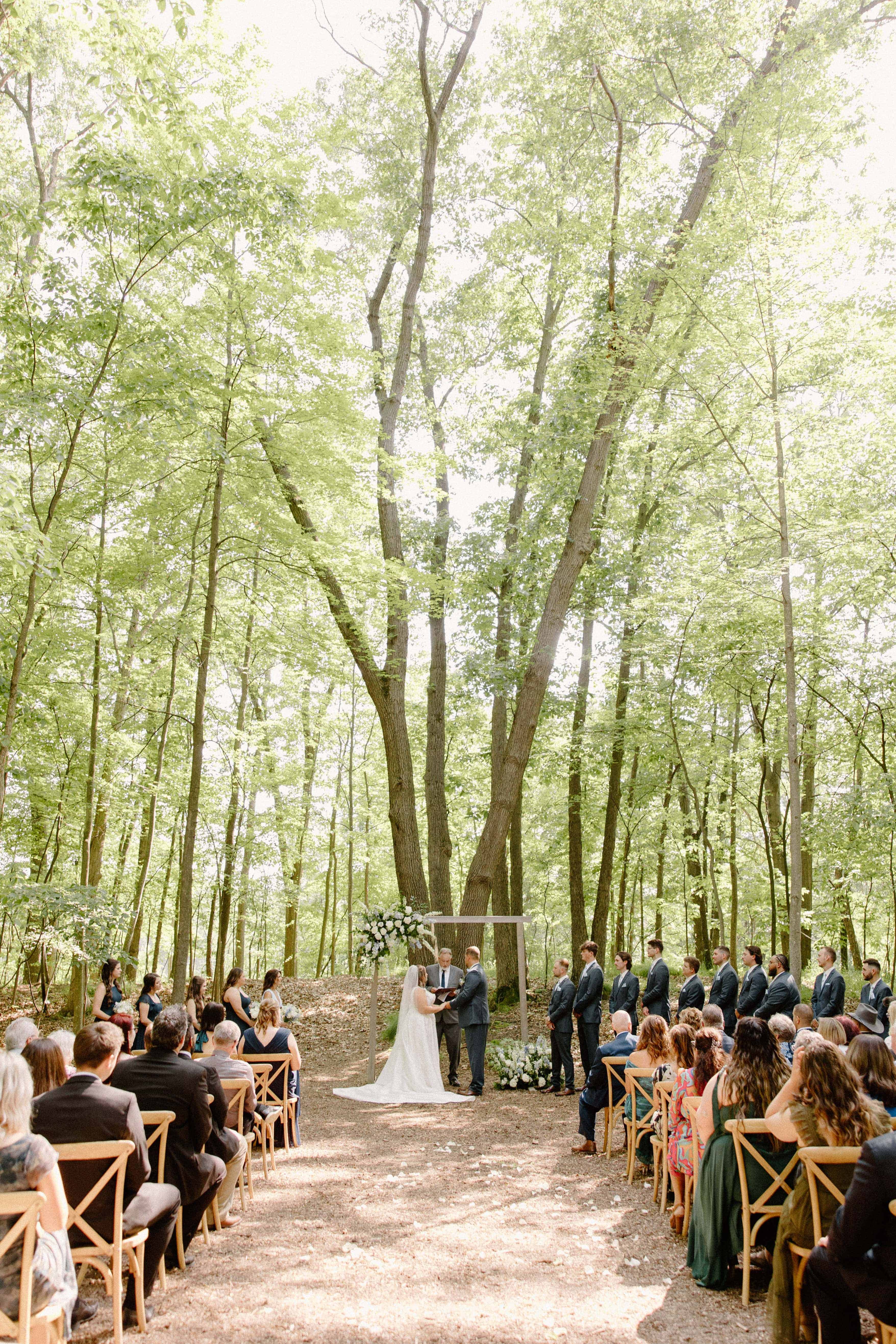  A wide-angle photo of a couple walking down a brick aisle toward the camera, surrounded by tall trees and seated wedding guests.