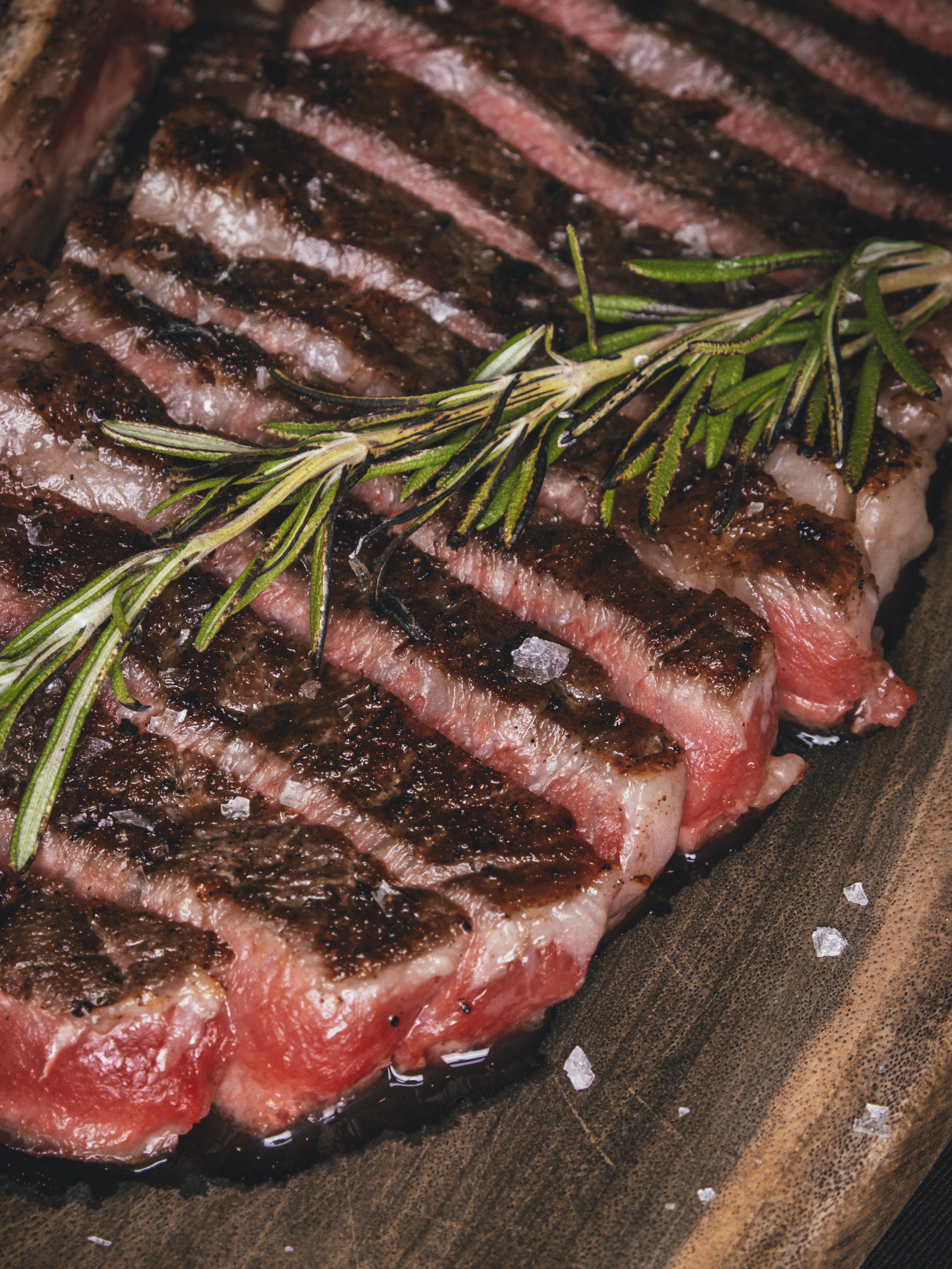 Close-up of a perfectly grilled steak sliced into thick pieces, topped with fresh rosemary and sprinkled with coarse salt, resting on a wooden platter.