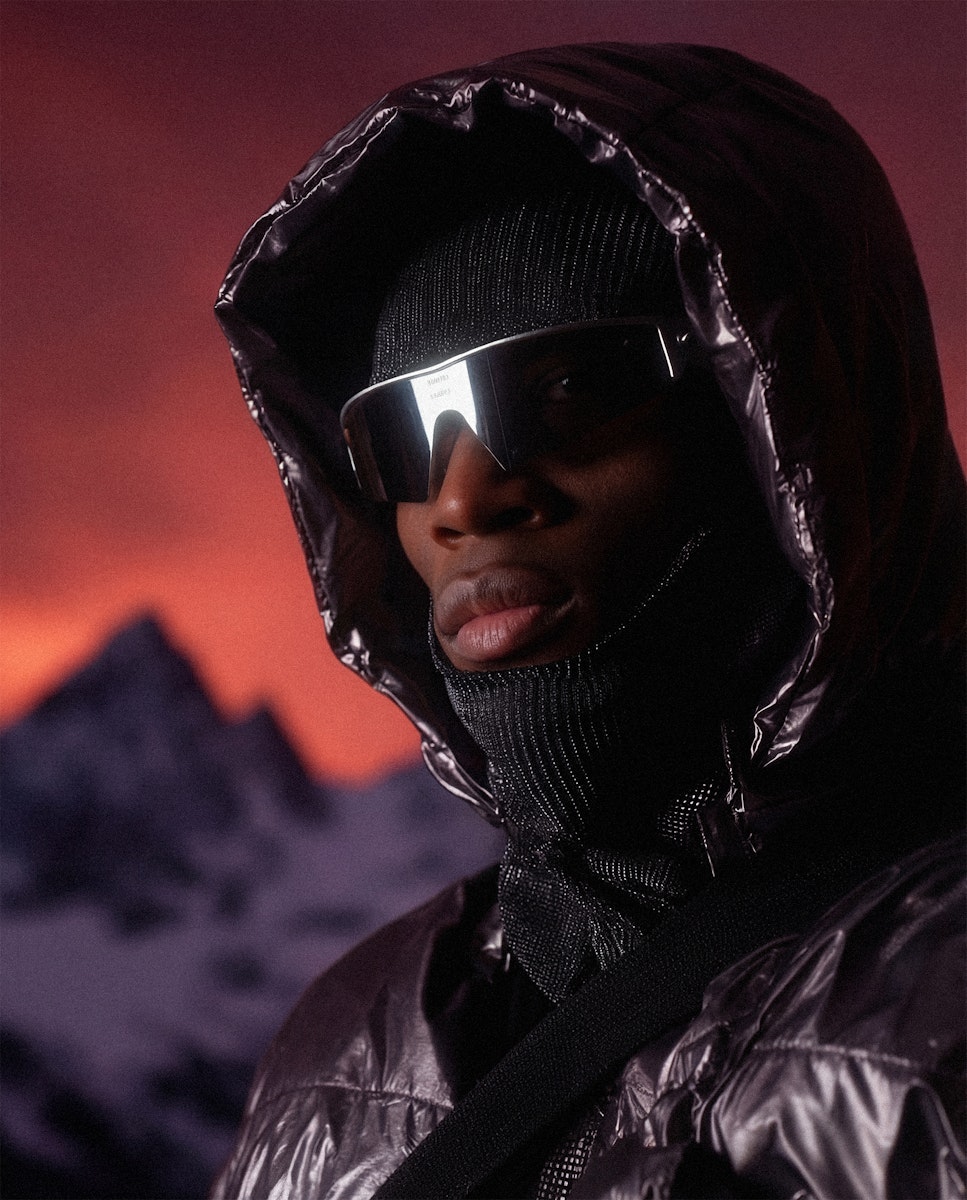 Portrait of a person in high-tech winter gear including a metallic hooded jacket, face mask, and futuristic sunglasses with mountains in the background.