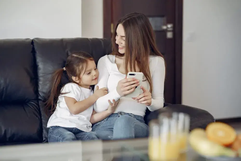 A smiling mother and daughter sit together on a black leather couch, sharing a joyful moment while looking at a smartphone. The image represents comfort, connection, and confidence in learning about home loan options with Chris Lewis Home Loans.