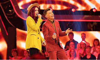 Two singers on a stage with a live audience. One in a yellow blazer and the other in a red suit, both holding microphones.