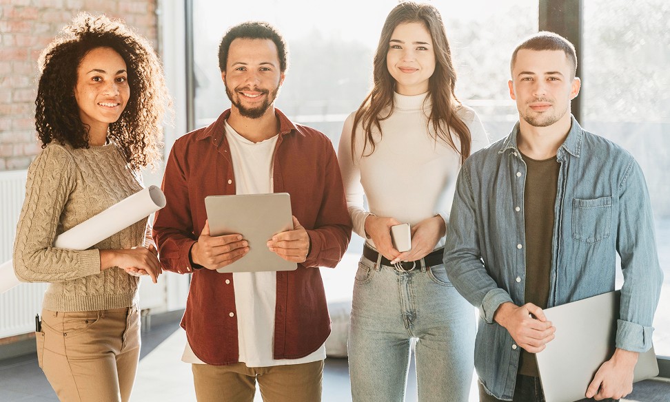 A diverse group of four people stands together, smiling, holding notebooks in an indoor setting.