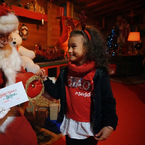 Ein Kind mit Rentiergeweih lächelt den Weihnachtsmann an, während es in einer festlichen Innenumgebung ein rotes Säckchen und einen Brief erhält.